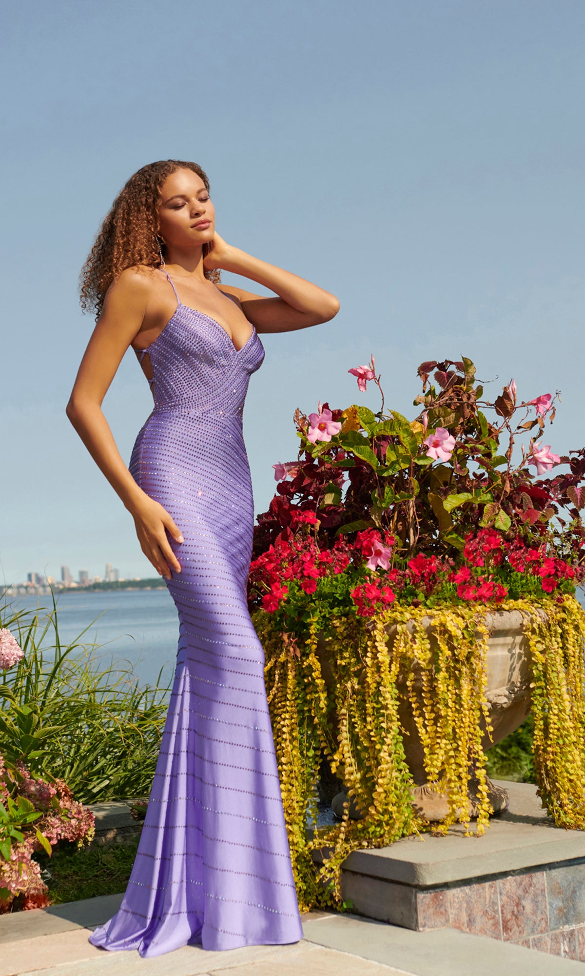 A woman in a fitted Backless Beaded Stretch Formal Dress by Faviana stands outdoors near pink flowers and greenery, with water and a city skyline in the background.